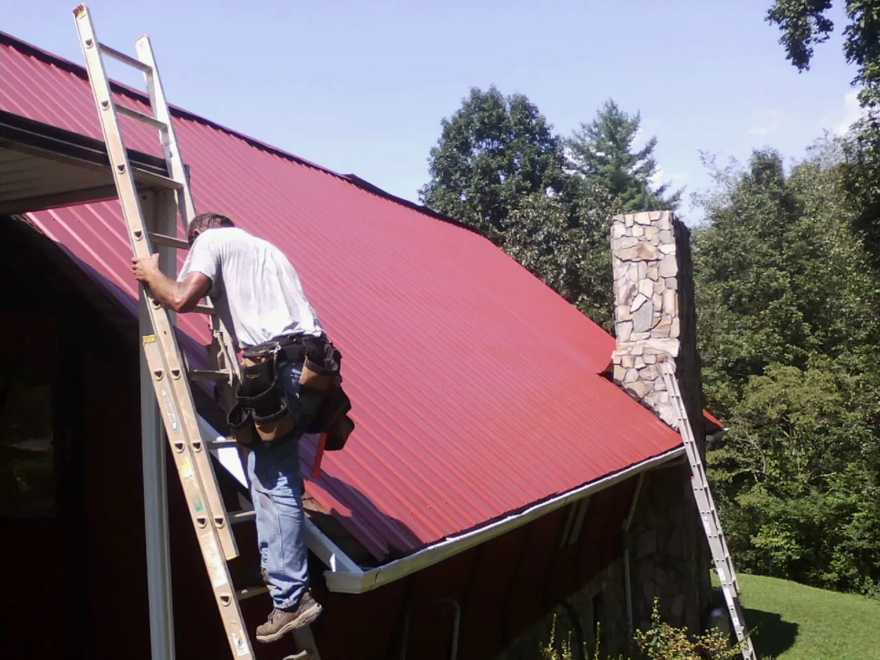 Roofer on a ladder beside a red metal roof and stone chimney — Soffit & Fascia Repair by Roofing Brothers in Maysville